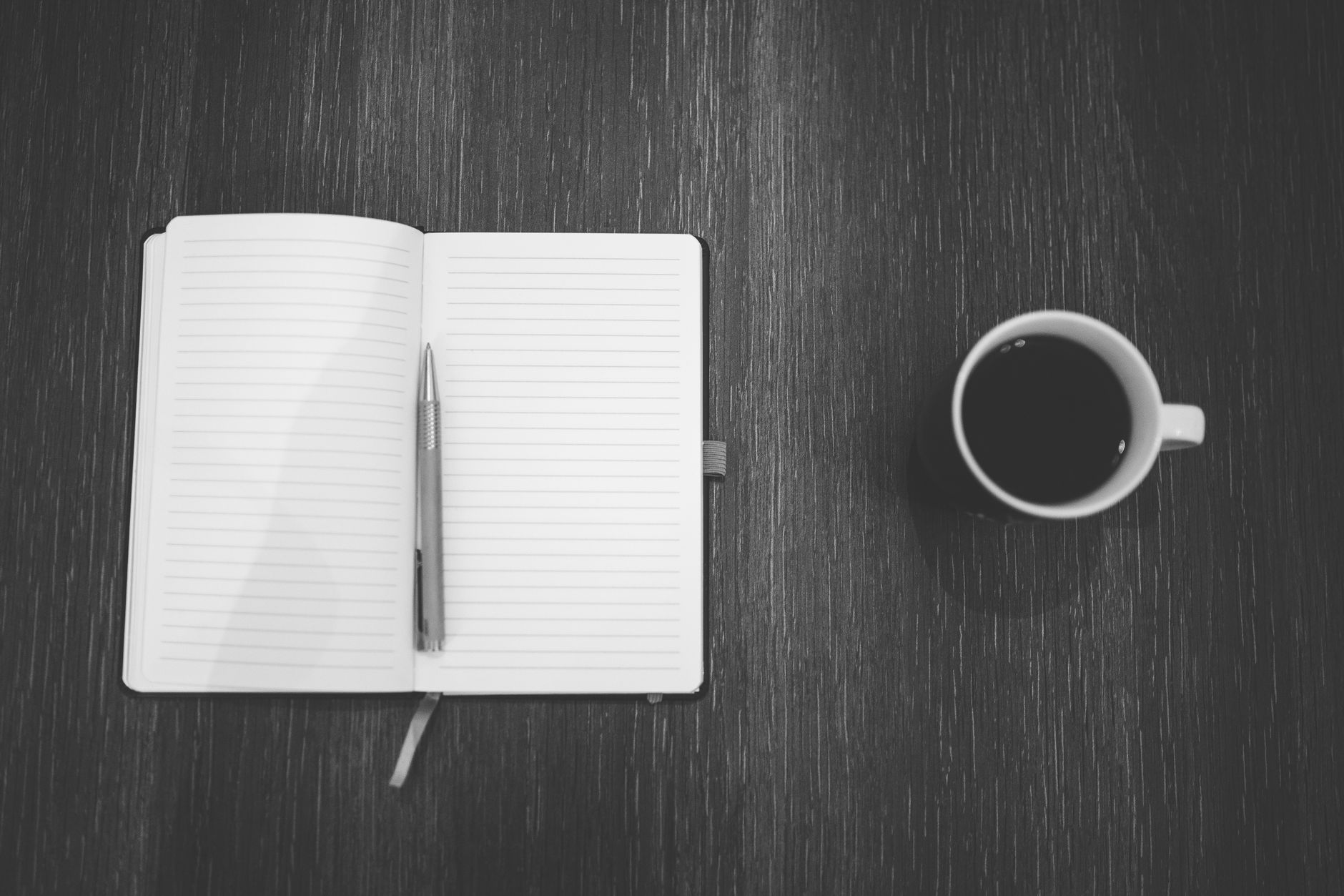 Black and white shot of a notepad and coffee cup on a wooden table from above.