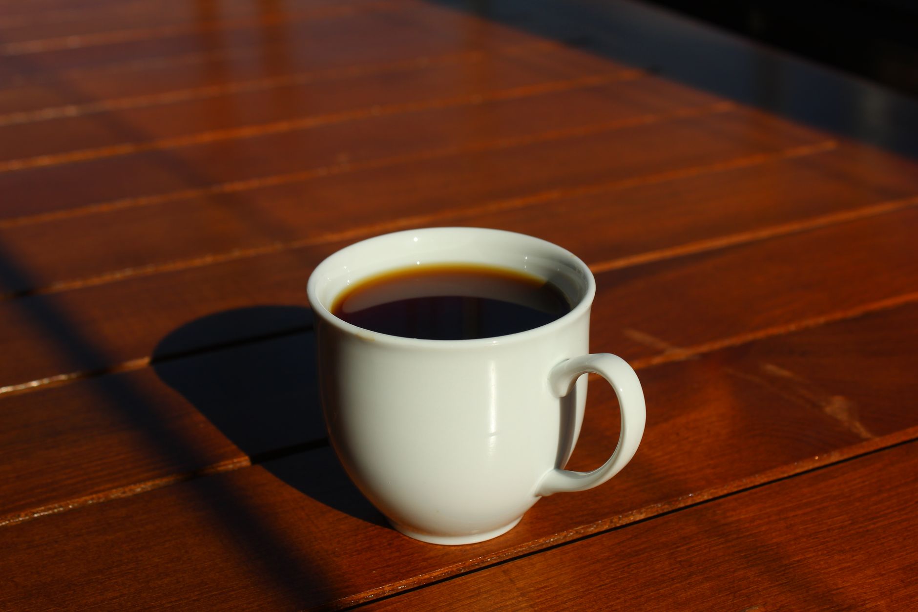A white coffee cup filled with black coffee on a wooden table in bright sunlight.