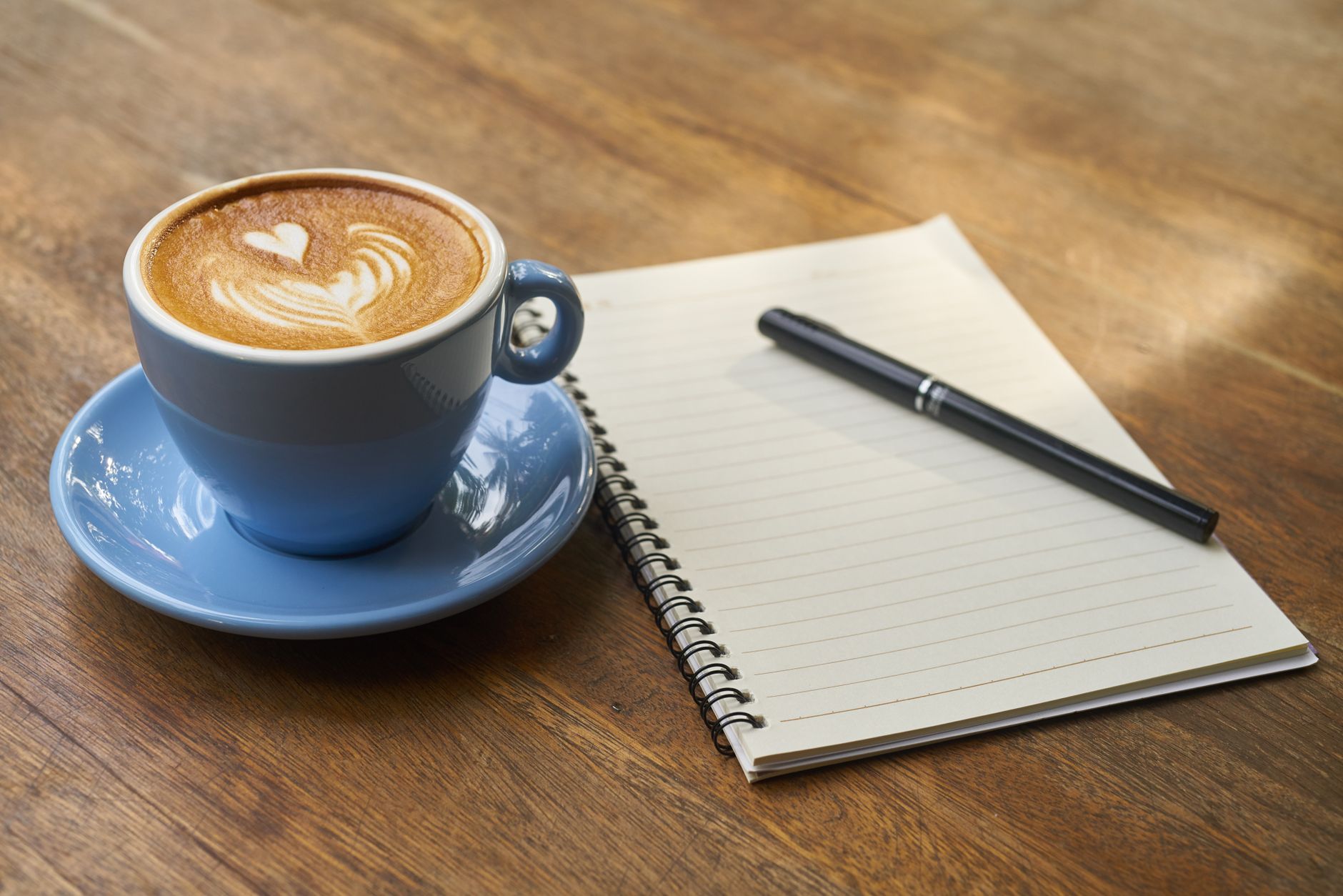 A calming scene of a latte with art in a blue cup beside a notebook and pen on a wooden table.