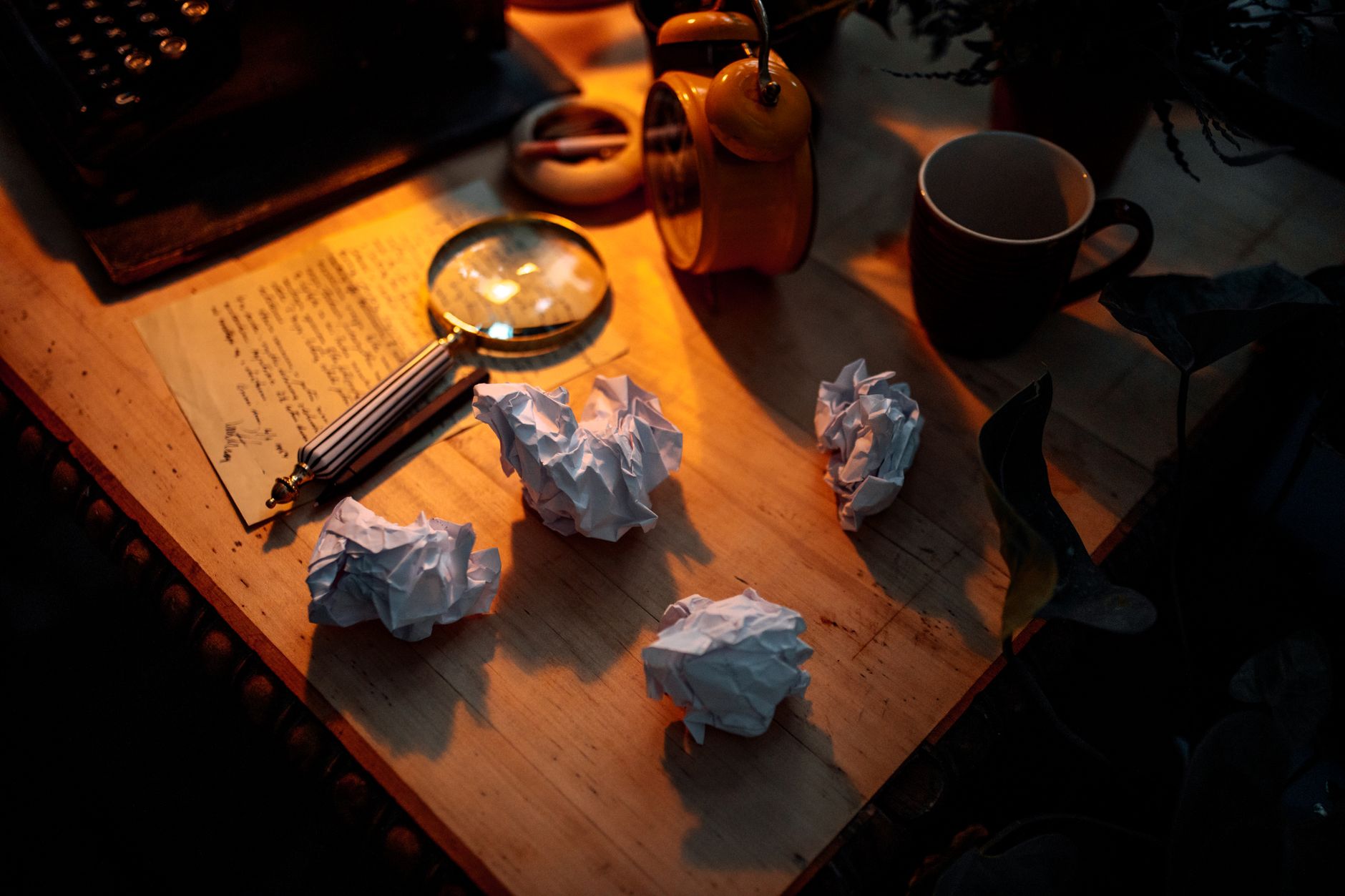 Ambient desk scene with crumpled papers, magnifying glass, and warm lighting setting a thoughtful mood.