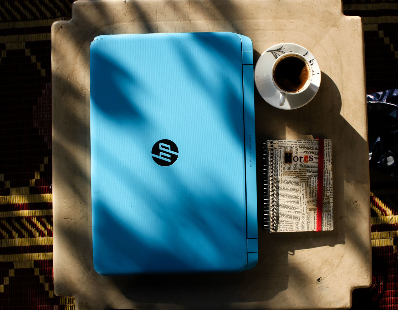 Flat lay of a blue laptop, coffee, and notebook on a table with shadows.