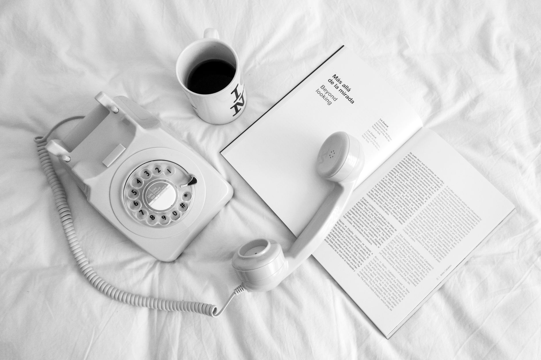 A cozy scene with a vintage rotary phone, coffee, and book on a white bed.