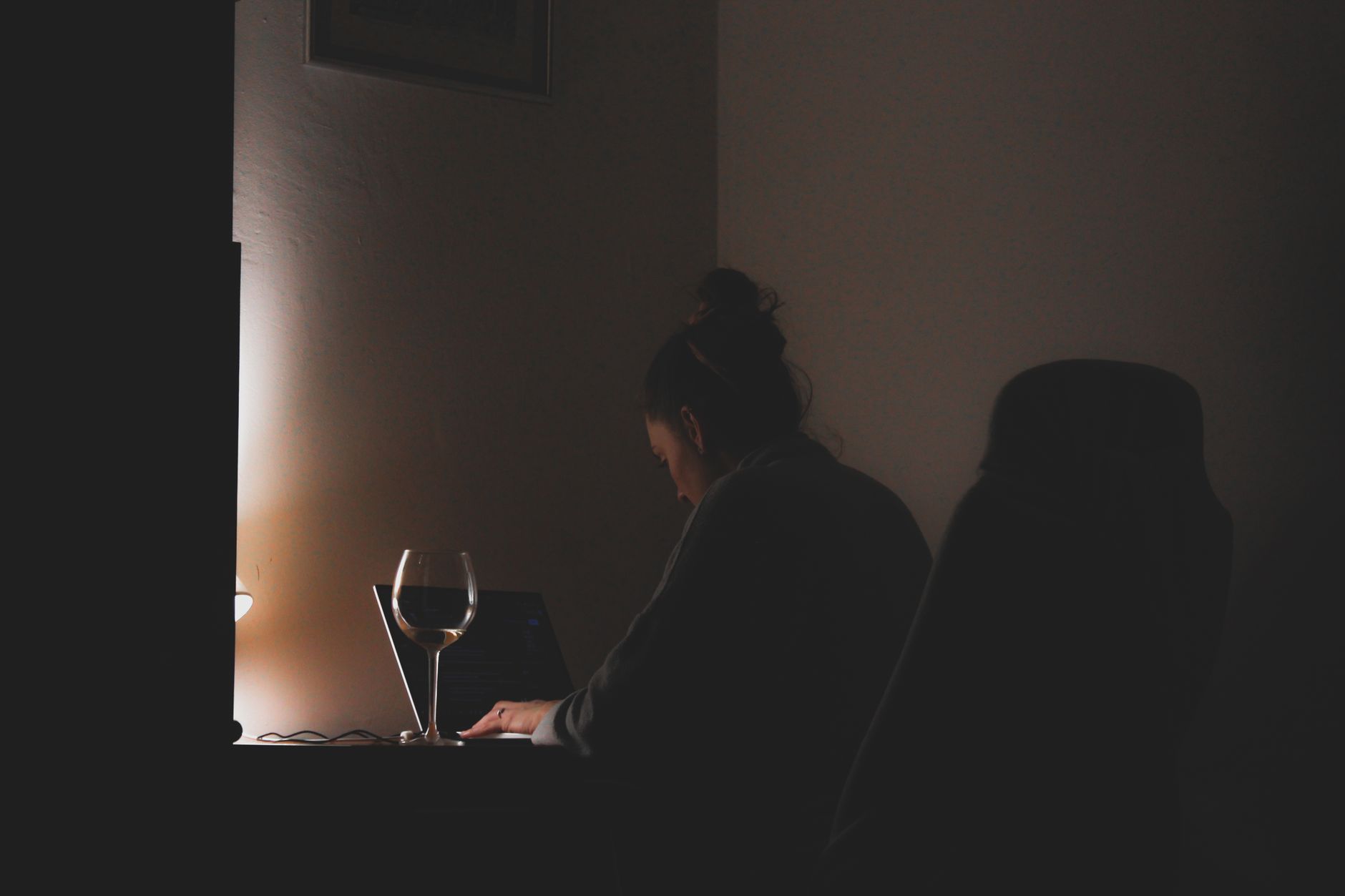 A woman working on a laptop with a glass of wine in a dimly lit room.
