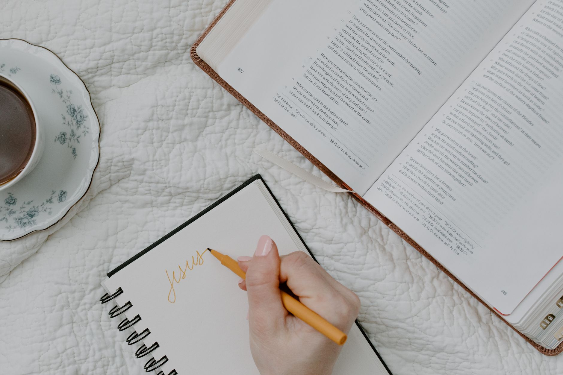A hand writing in a notebook next to an open Bible and a cup of coffee on a quilted blanket.