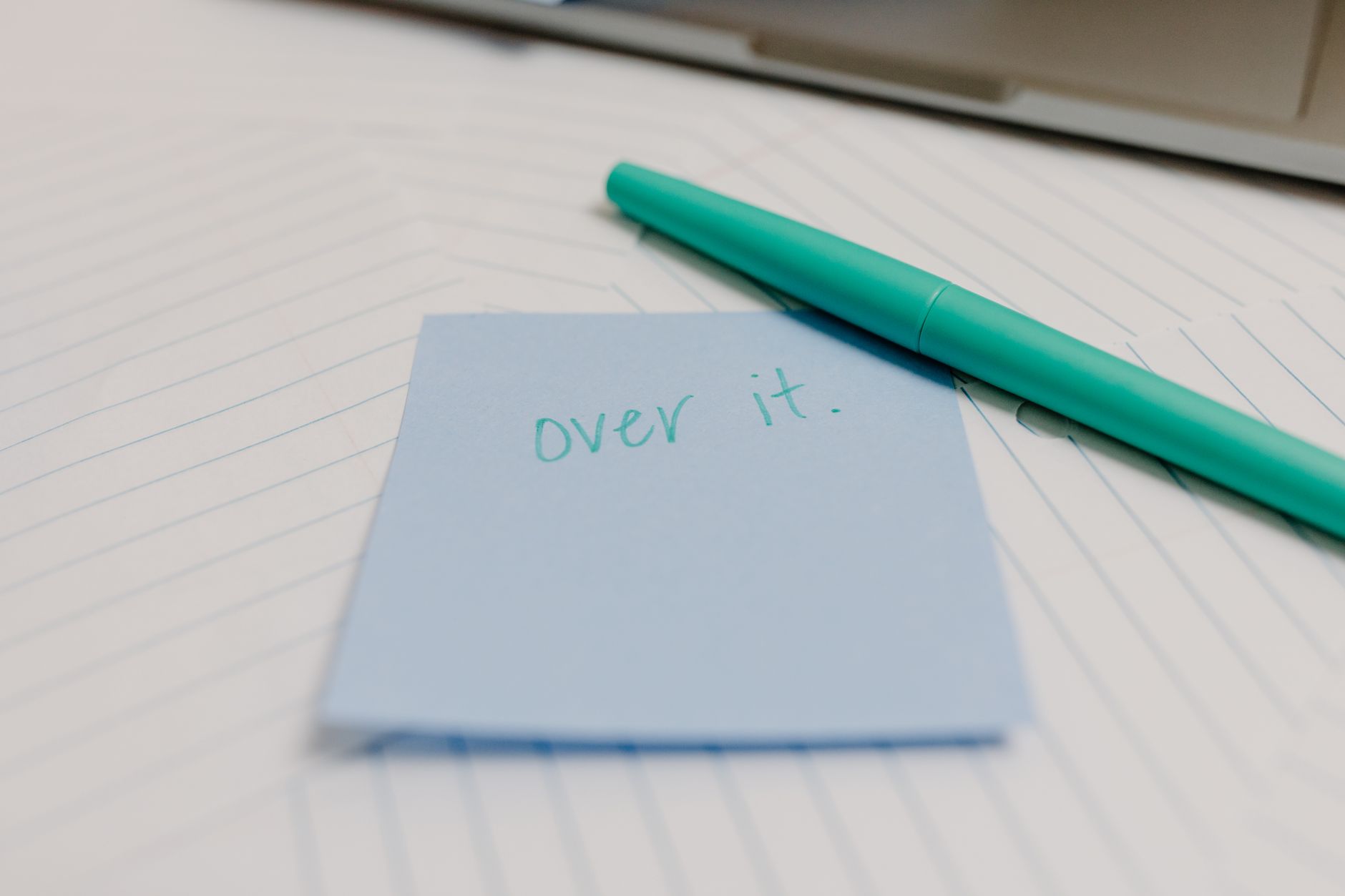 A blue sticky note with the message 'over it' beside a teal pen on lined paper.