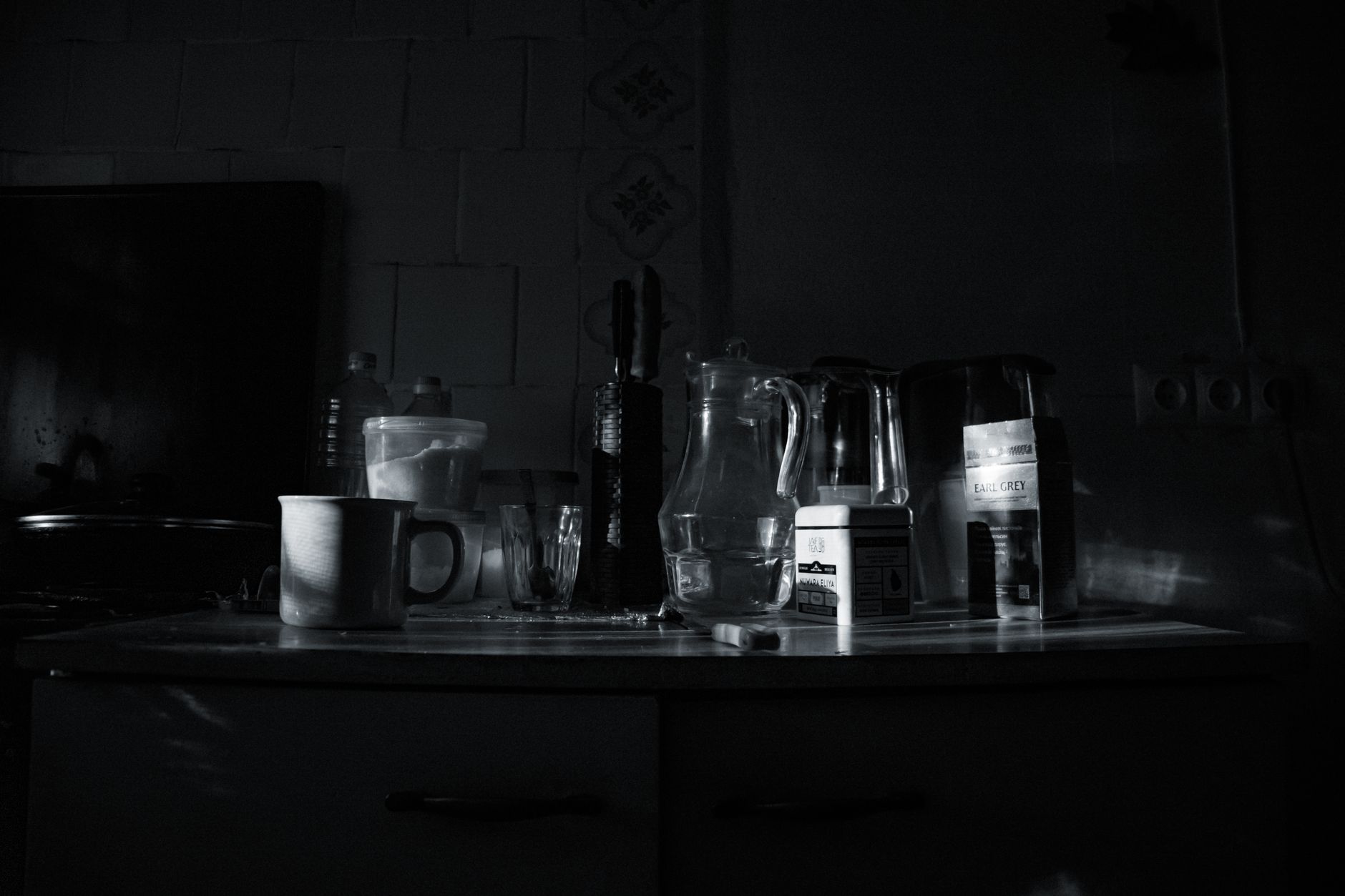 A moody kitchen scene featuring glassware, mugs, and containers in monochrome lighting.
