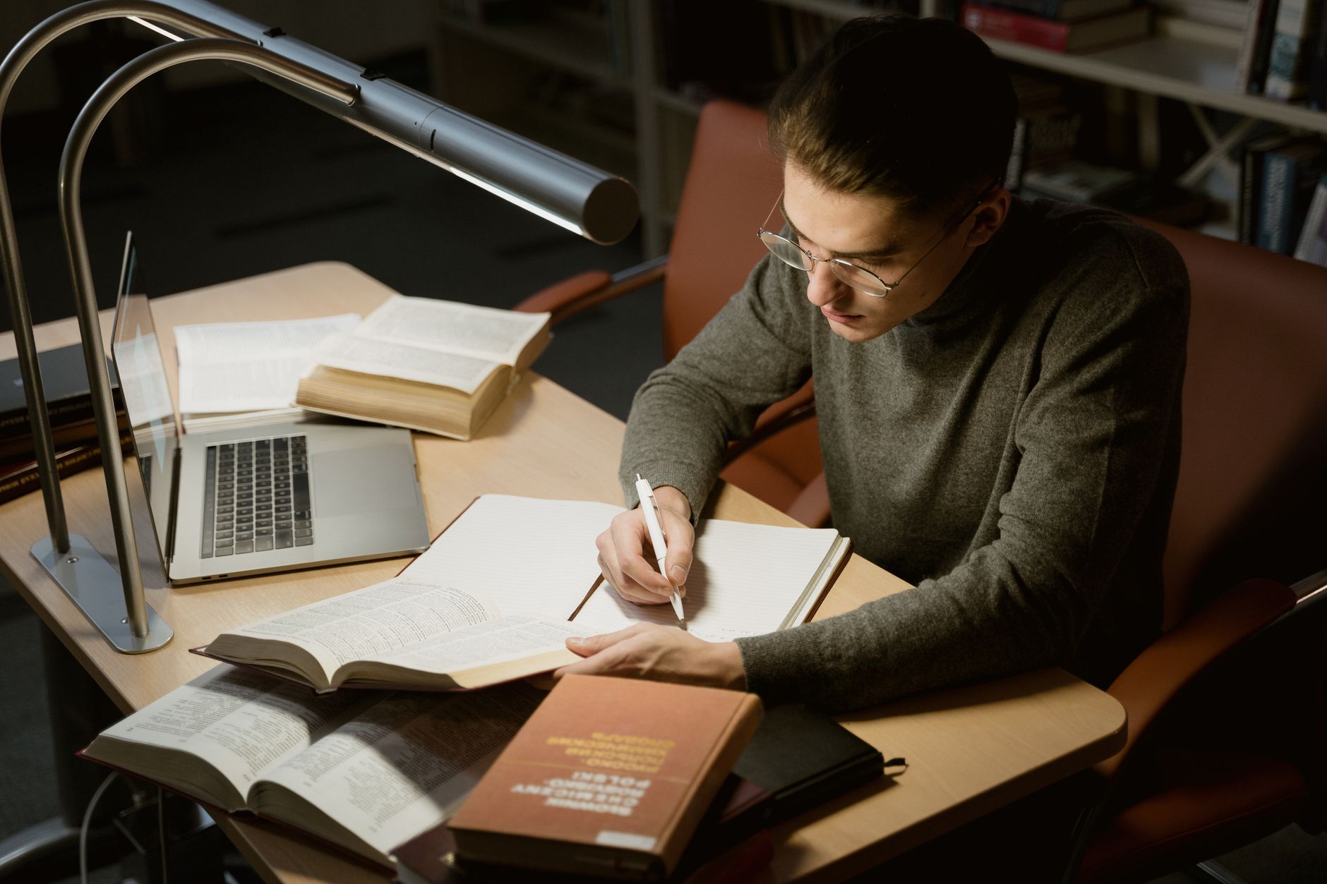 A young adult studying with books and a laptop, focused on taking notes at a desk under a lamp.