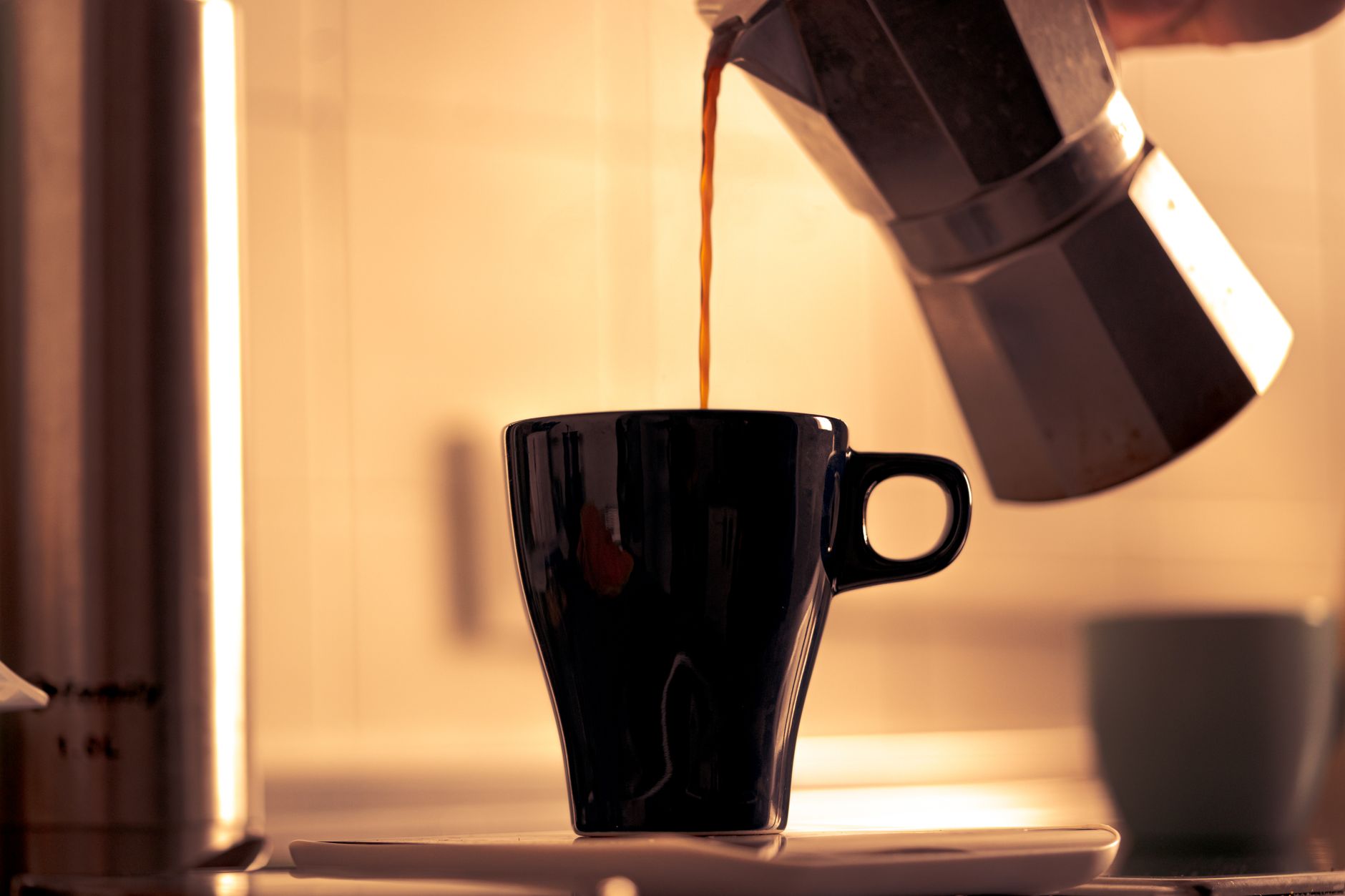 Close-up of fresh coffee being poured into a ceramic cup from a moka pot in a warmly lit kitchen.