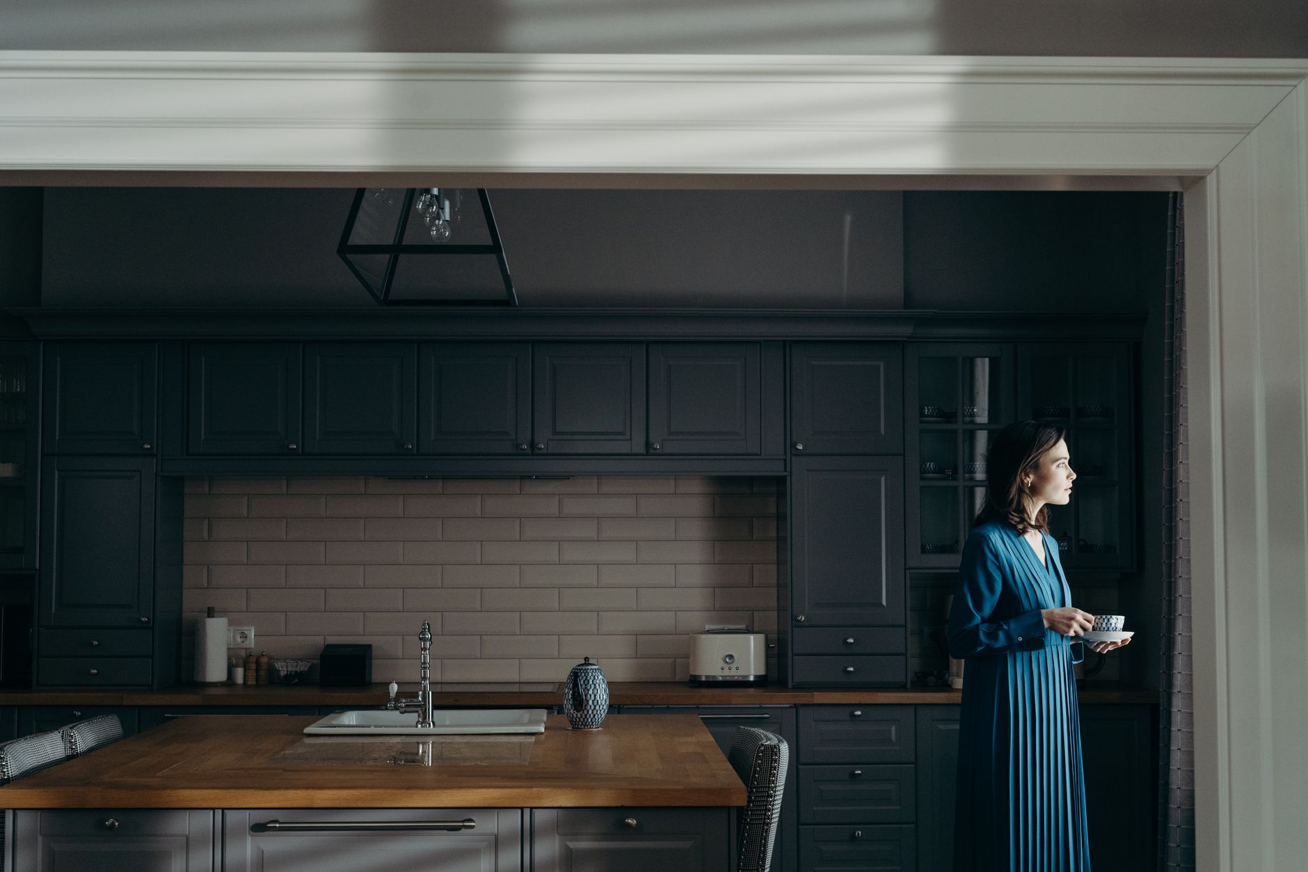 A woman in a blue dress stands in a kitchen holding a cup and saucer, bathed in natural light.