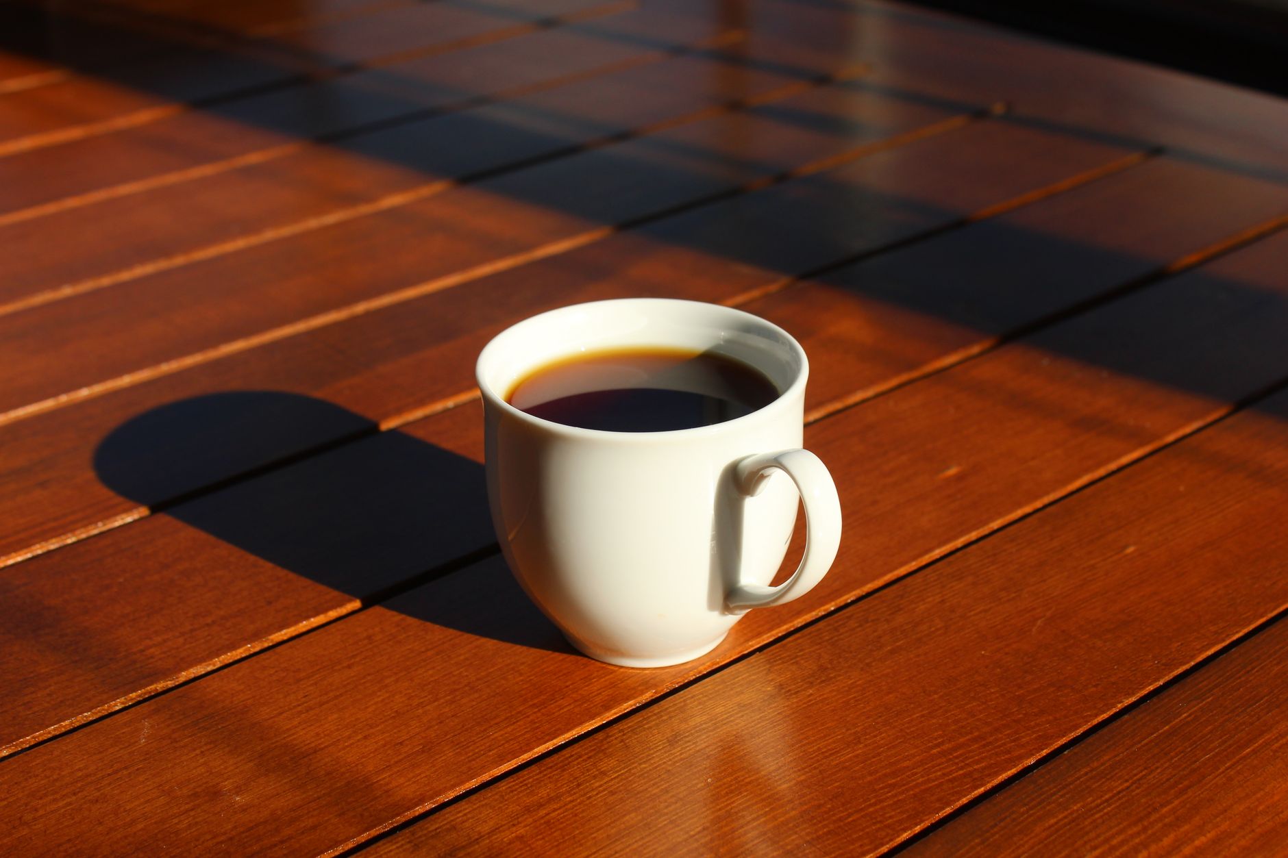 A white coffee cup sits in morning sunlight on a wooden table casting a shadow.