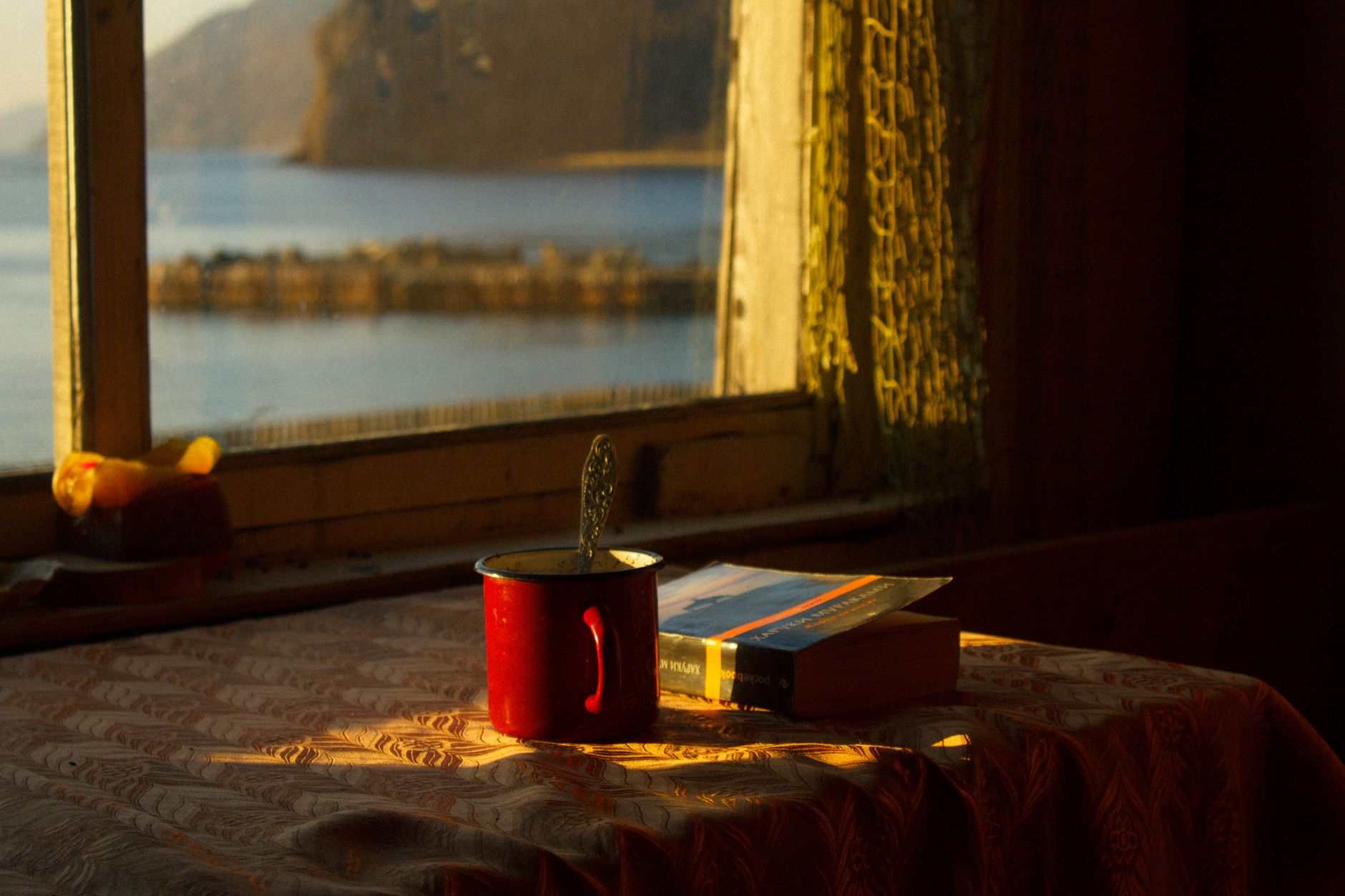 A peaceful scene with a red cup and book by a lake-view window, perfect for cozy reading moments.
