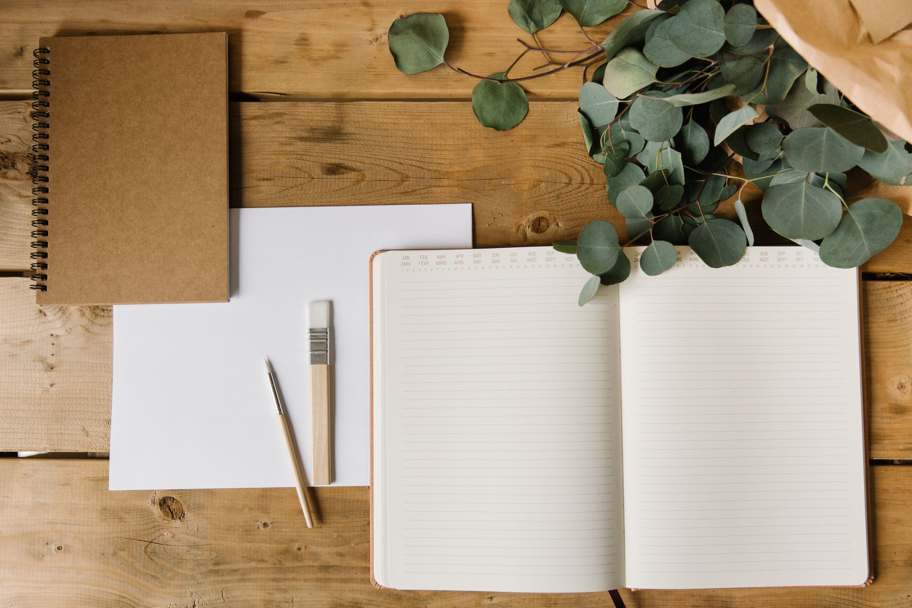 Elegantly styled flat lay featuring notebooks, eucalyptus leaves, and writing tools on a wooden surface.