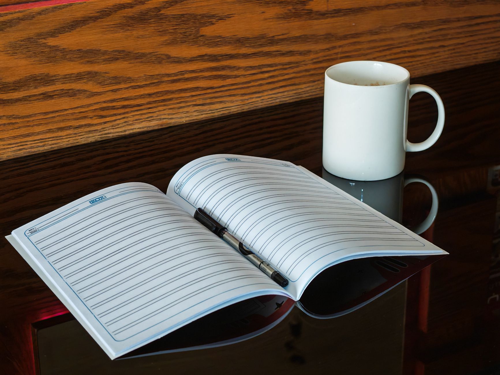 An open notebook with a pen beside a coffee mug on a reflective table.
