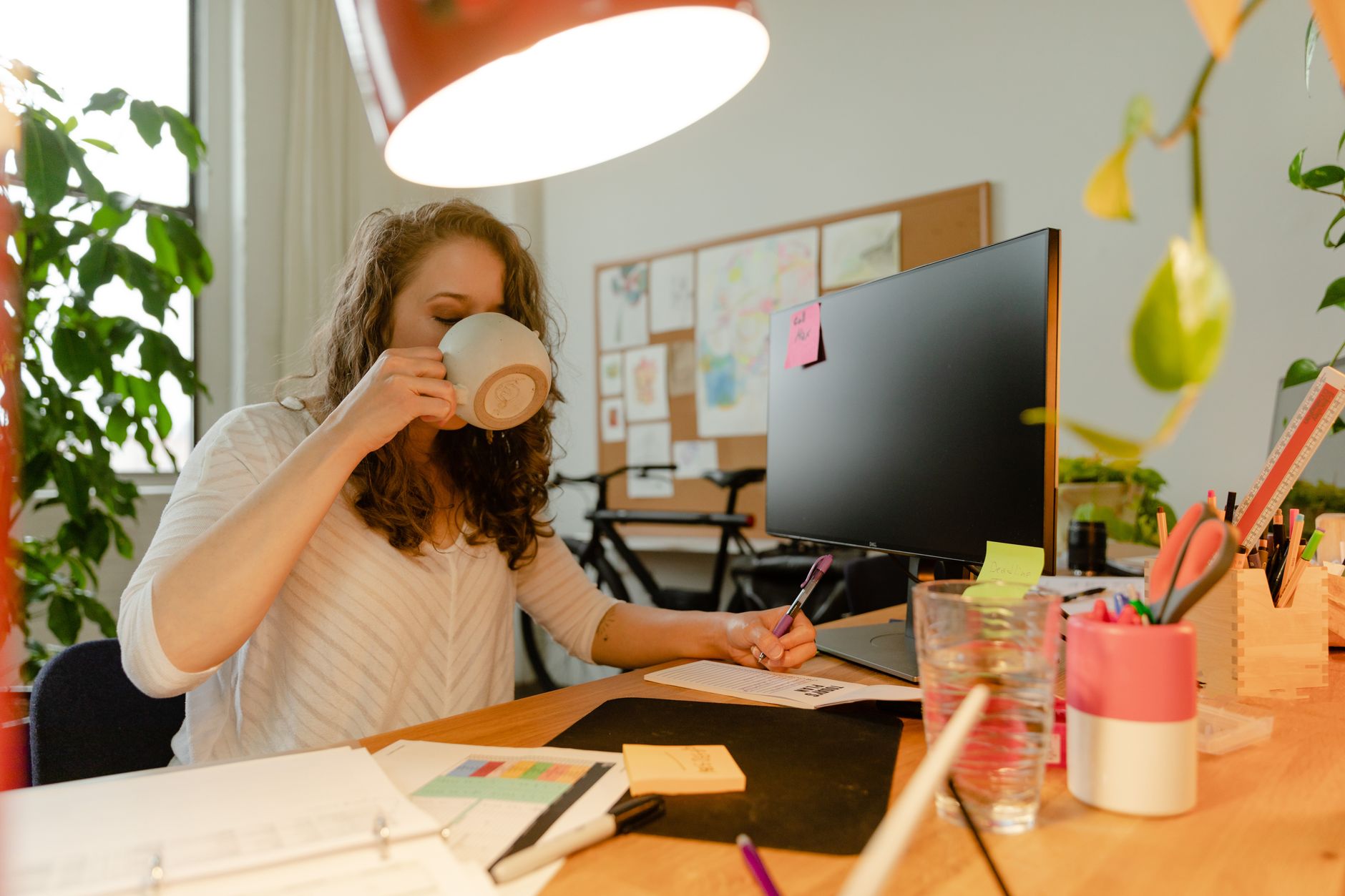 Professional woman drinking coffee at a desk in a modern office environment with plants.