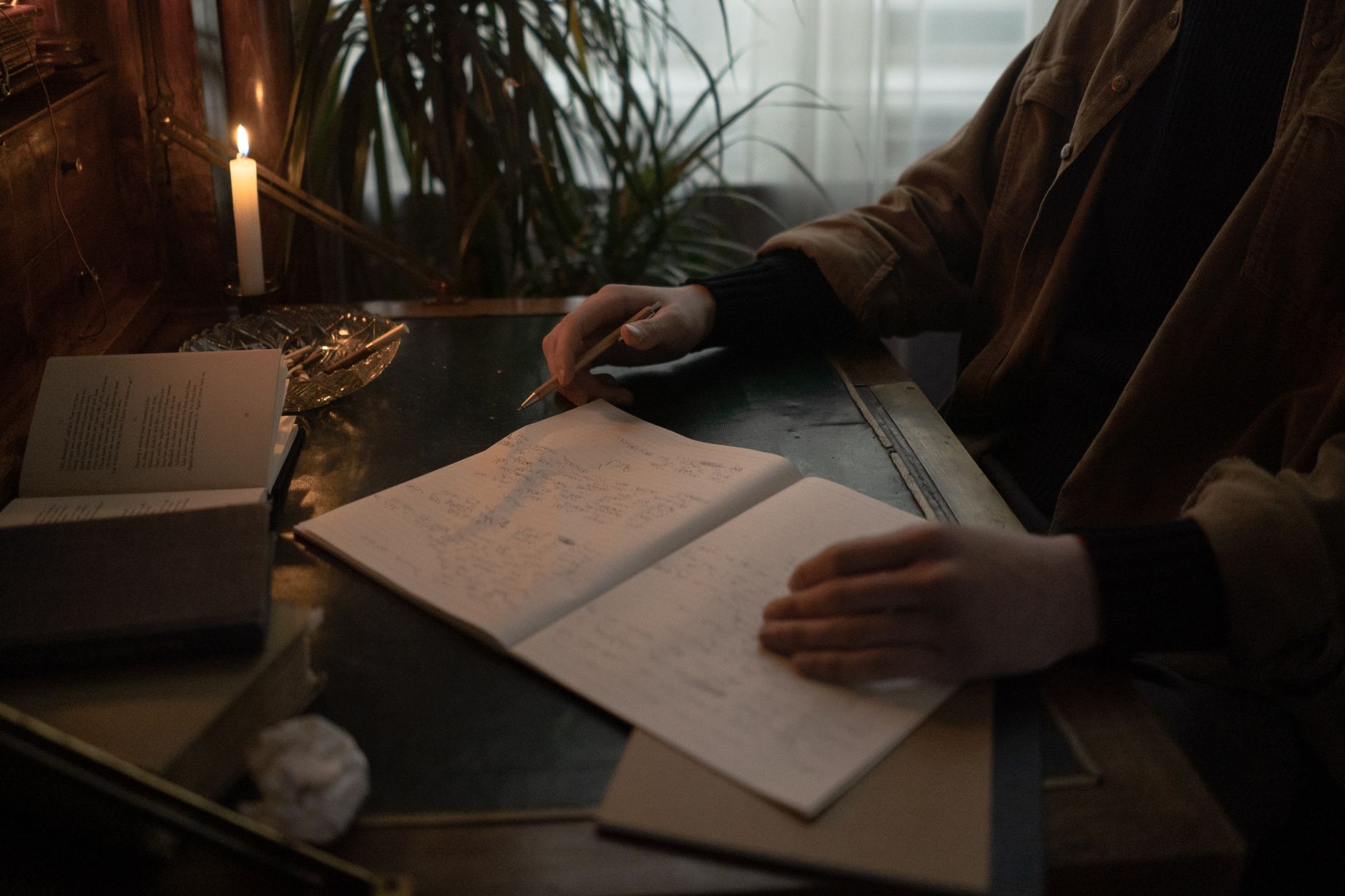 A person writes in a dimly lit room, surrounded by open books and candlelight, creating a moody study atmosphere.