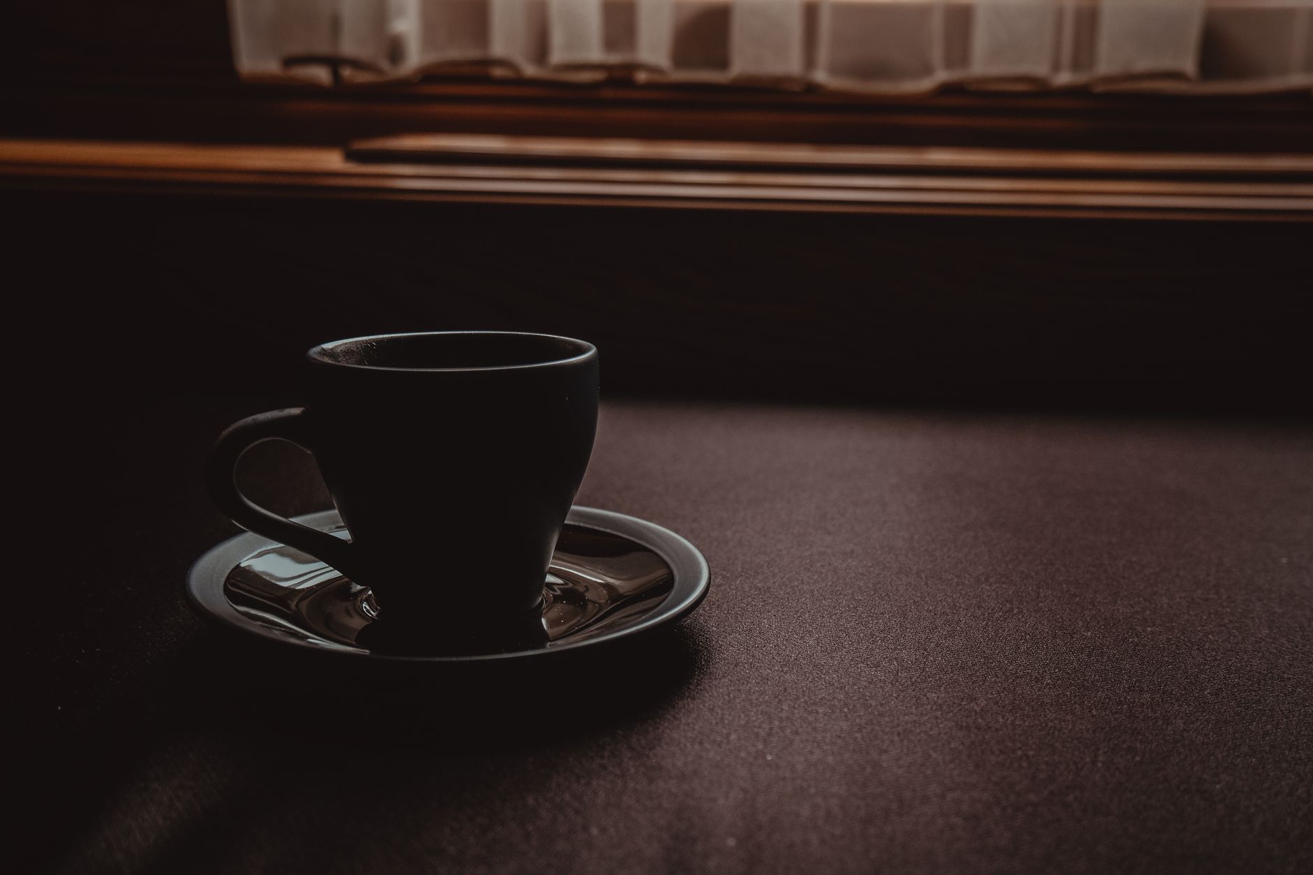 A minimalistic dark-toned image featuring a coffee cup on a table near a window.