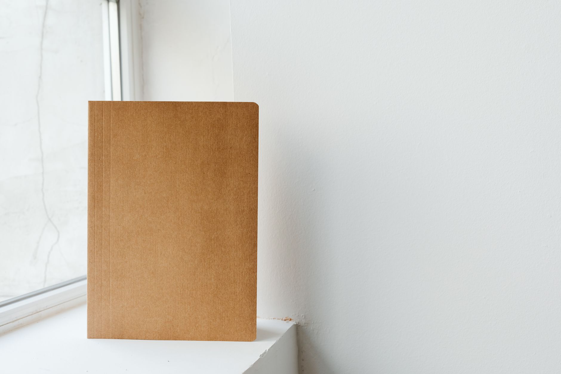 Minimalist photo of a brown notebook placed against a white wall near a window, offering plenty of copy space.