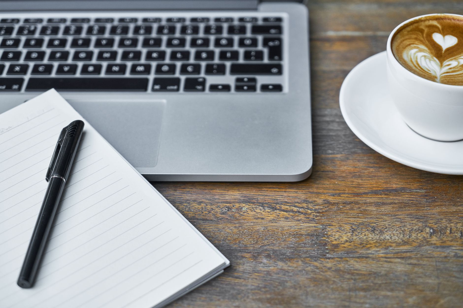 A cozy workspace featuring a laptop, notebook, and coffee cup with latte art on a rustic table.
